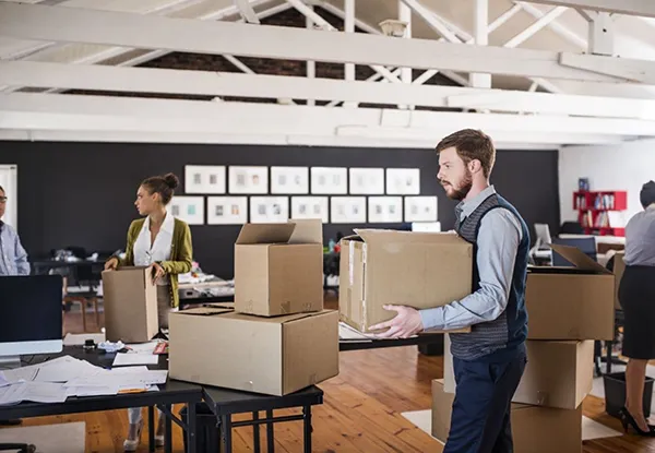 Two movers unpack cardboard boxes in a bright office space, with desks and office equipment visible in the background.