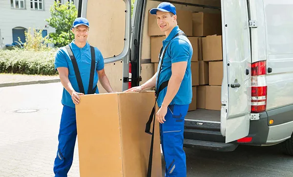 Two movers in blue uniforms carefully maneuver a large cardboard box out of the back of a white moving van.