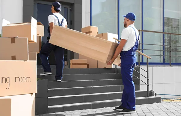 Two movers in uniform carefully carry a long, heavy wooden piece of furniture up a short set of stairs and into a moving truck.