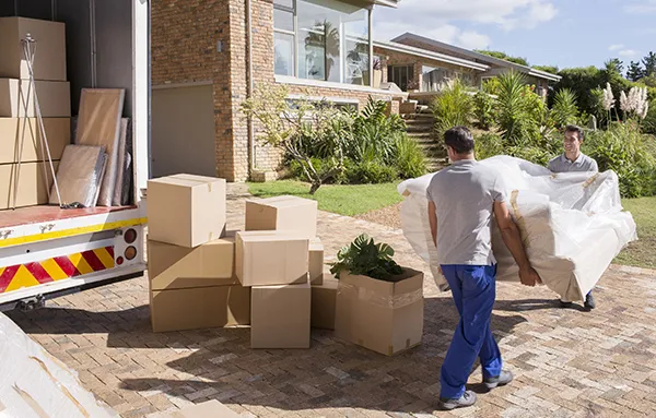 Two movers use a dolly to move a stack of cardboard boxes towards a residential house on a sunny day.