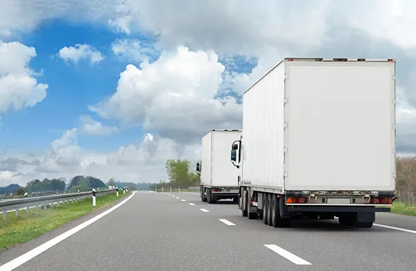 Two large white moving trucks drive down a highway on a sunny day, passing green fields.