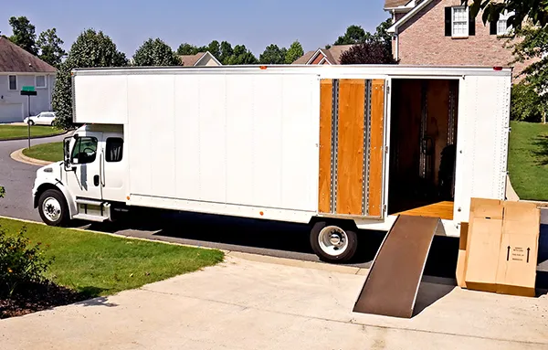 A white moving truck is parked on a street with its back door open, revealing stacks of cardboard boxes inside. Several more boxes are on the ground next to the truck.