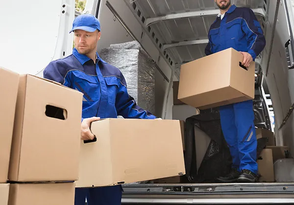 A mover in a blue uniform carries a cardboard box out of the back of a moving truck, while another mover is inside the truck.